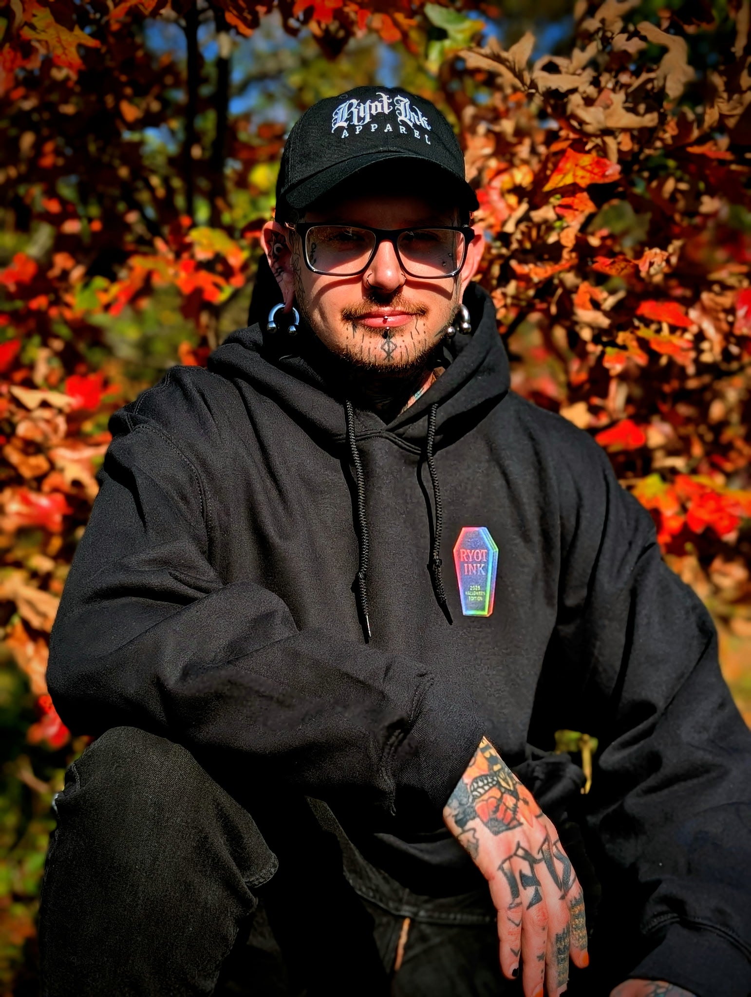 Person wearing a black hoodie and cap with tattoos, sitting among autumn leaves.